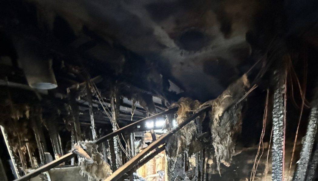 Severely fire-damaged interior with charred wooden framing, hanging burnt insulation, and soot-blackened ceiling.