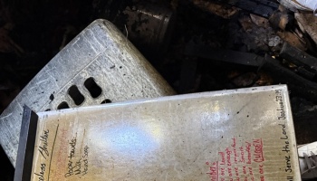 A soot-stained sign listing shelter supplies and handwritten encouraging messages sits among charred fire debris.
