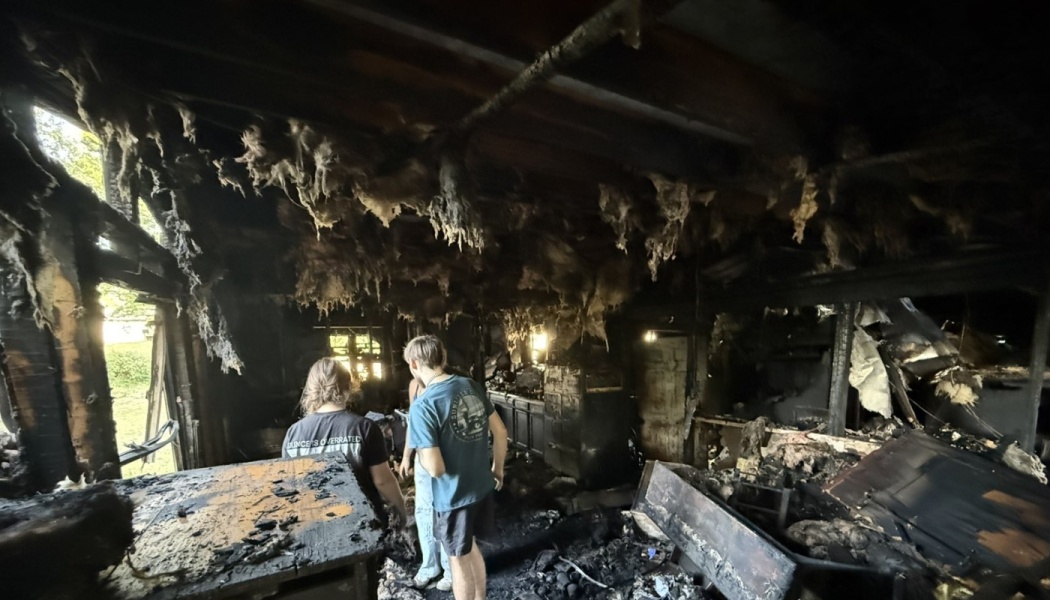 Two people inspect extensive fire damage inside a destroyed home with charred walls and burnt ceiling insulation.
