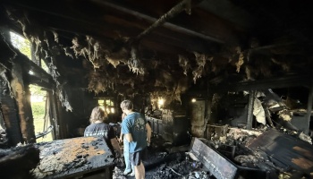 Two people inspect extensive fire damage inside a destroyed home with charred walls and burnt ceiling insulation.