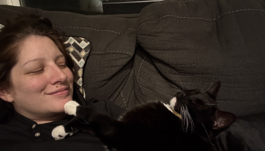 A smiling woman lies on a dark couch, cuddling with a black and white cat that rests its paw on her chin.