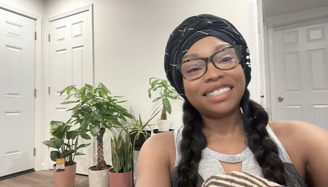 A smiling woman with glasses and a black headwrap sitting in front of several potted houseplants.