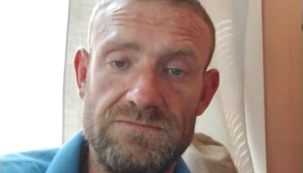 Close-up portrait of a man with short hair and a scruffy beard, wearing a bright blue collared shirt.