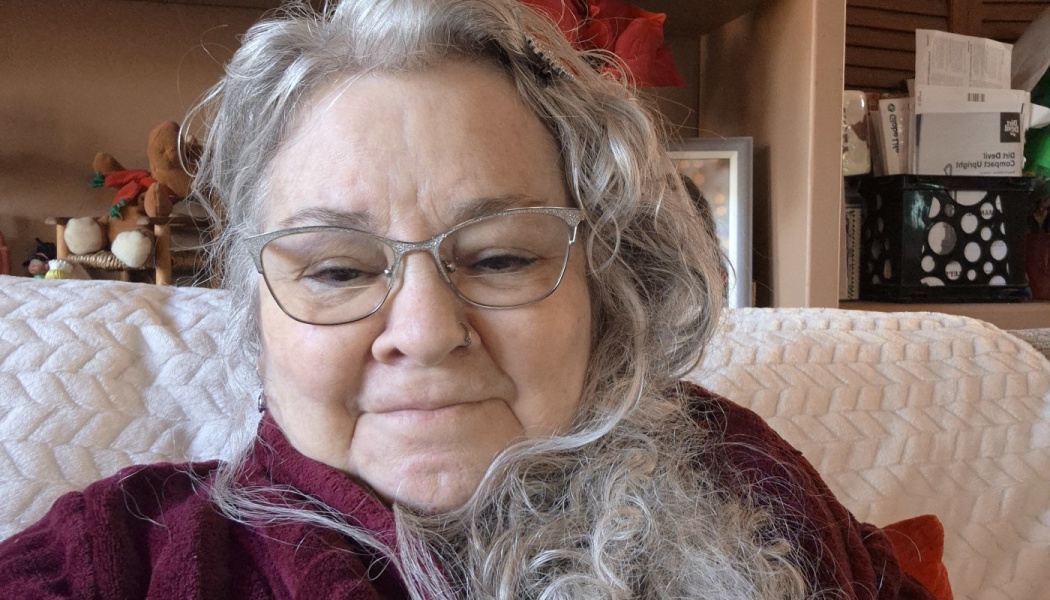 An older woman with long, wavy grey hair and sparkly cat-eye glasses smiling softly in a maroon sweater.