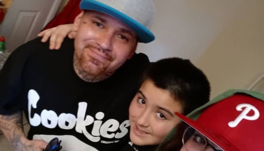 A smiling tattooed man wearing a blue cap and Cookies t-shirt poses closely with a young boy.
