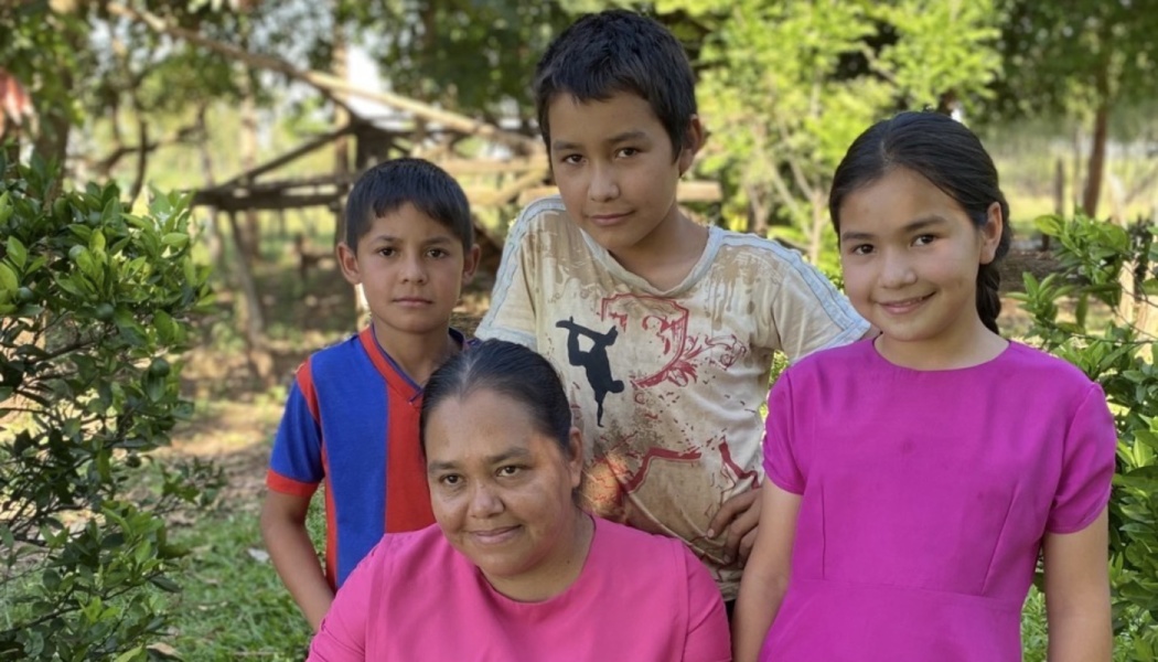 A smiling mother poses outdoors with her three young children surrounded by lush green foliage.