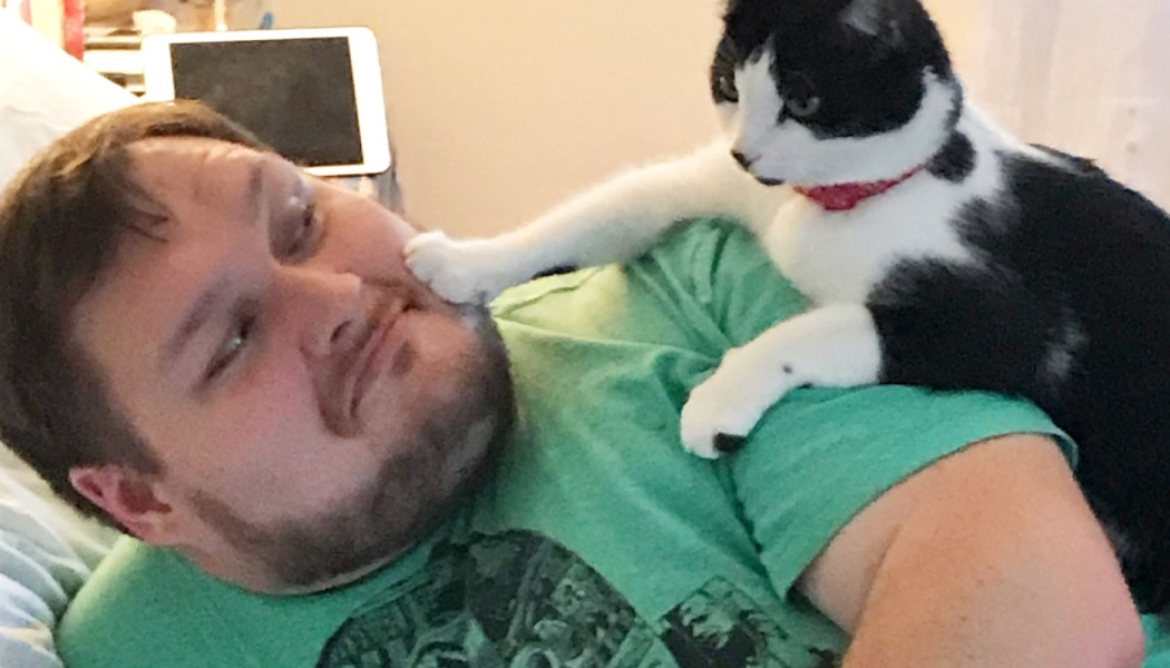 A black and white cat sitting on a man's chest places its paw gently on his cheek.