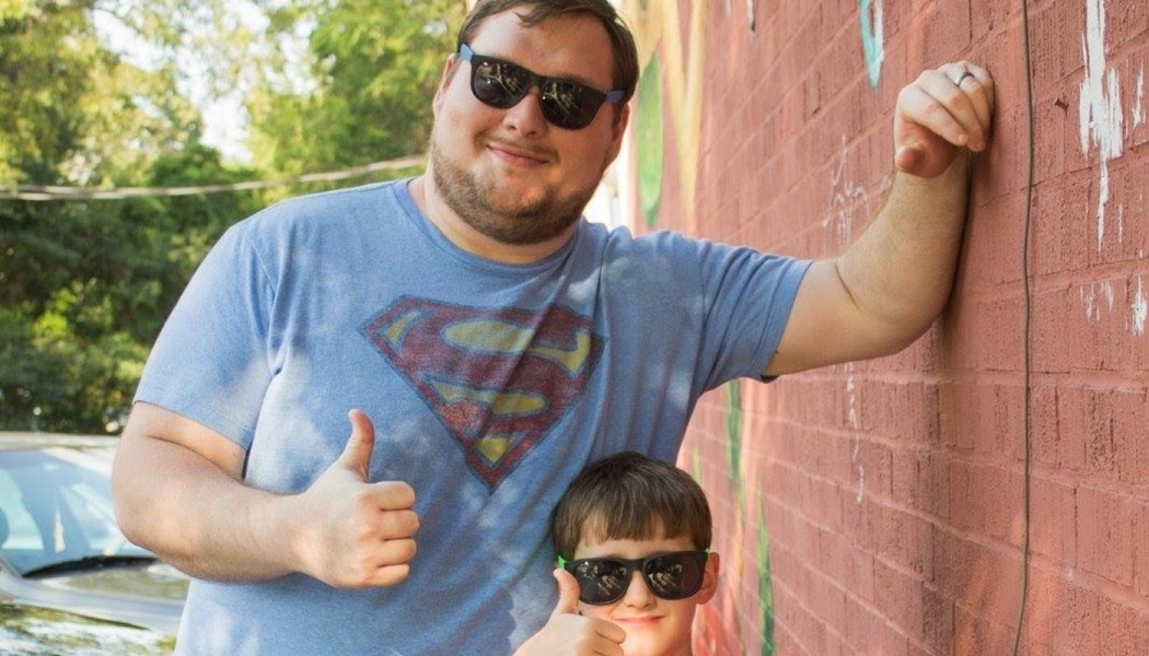 A man in a blue Superman shirt and a young boy, both wearing sunglasses and giving thumbs-up signs by a brick wall.