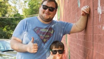 A man in a blue Superman shirt and a young boy, both wearing sunglasses and giving thumbs-up signs by a brick wall.