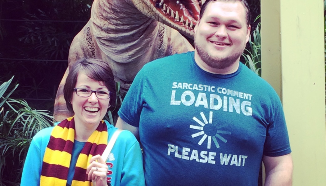 A smiling woman wearing a striped scarf and a man in a blue graphic tee pose together in front of a dinosaur statue.