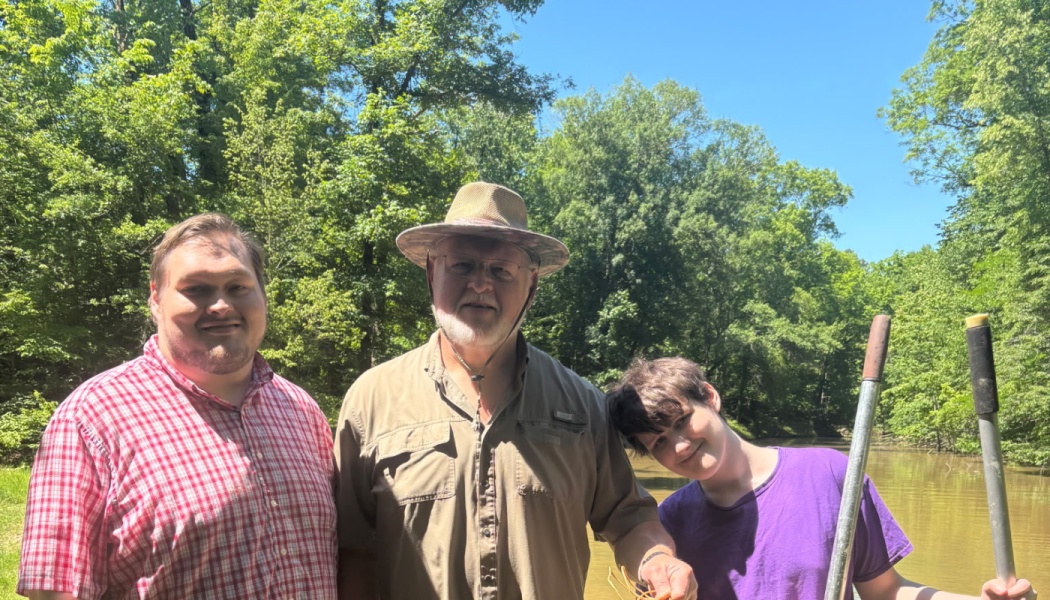 Three smiling men pose together on a boat along a tree-lined river on a bright, sunny day.
