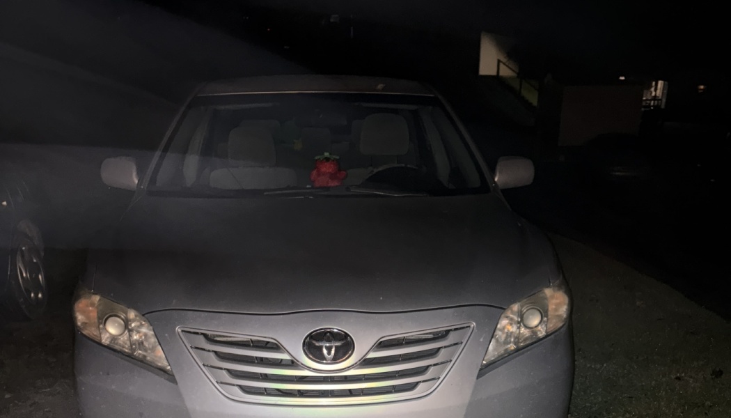 Front view of a silver Toyota sedan parked at night with a small red plush toy on the dashboard.