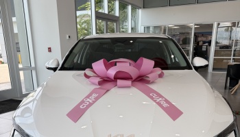 White car in a dealership with a large pink breast cancer awareness bow reading CURE on the hood.