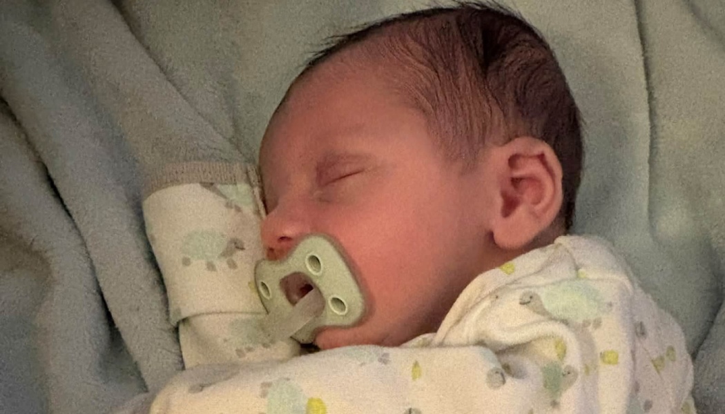 A sleeping newborn baby with dark hair using a pacifier, resting on a soft blanket.