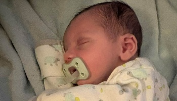 A sleeping newborn baby with dark hair using a pacifier, resting on a soft blanket.