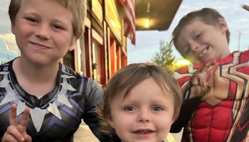 Three smiling young boys posing together outdoors, with two dressed in superhero costumes.