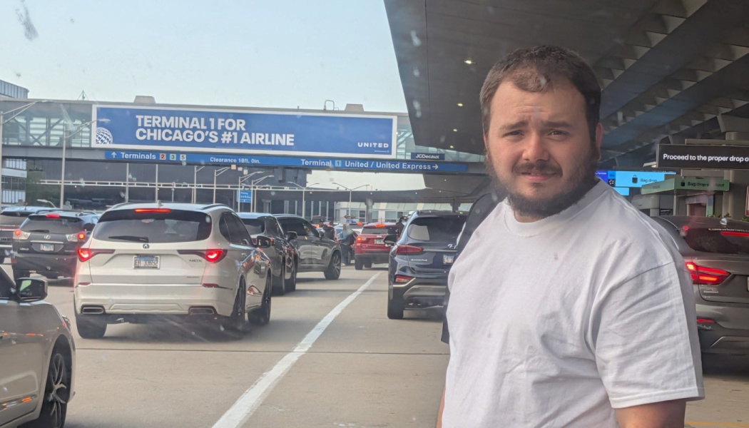 A bearded man in a white t-shirt stands outside a busy airport terminal drop-off zone with heavy traffic.
