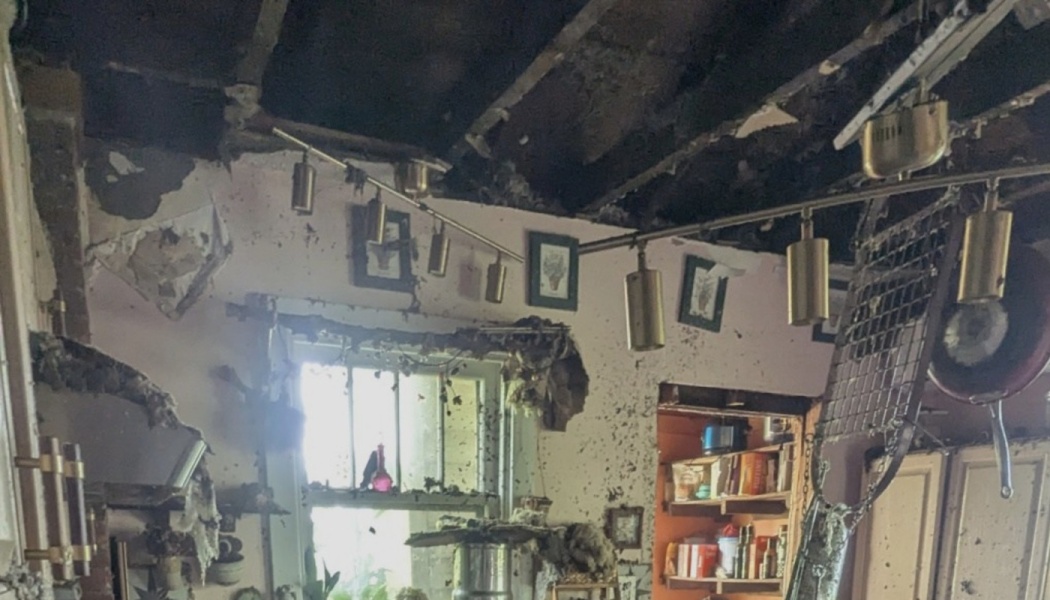Severely fire-damaged room with a charred ceiling, exposed rafter beams, soot-stained walls, and hanging lights.