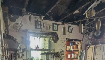 Severely fire-damaged room with a charred ceiling, exposed rafter beams, soot-stained walls, and hanging lights.