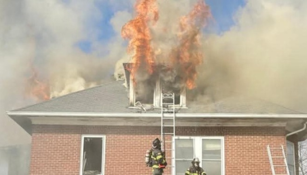 Flames and thick smoke burst from the roof of a red brick house while firefighters respond below.