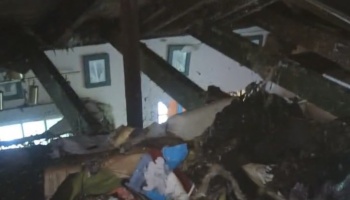 Severely fire-damaged home interior with charred ceiling beams, collapsed roof, and scattered debris.