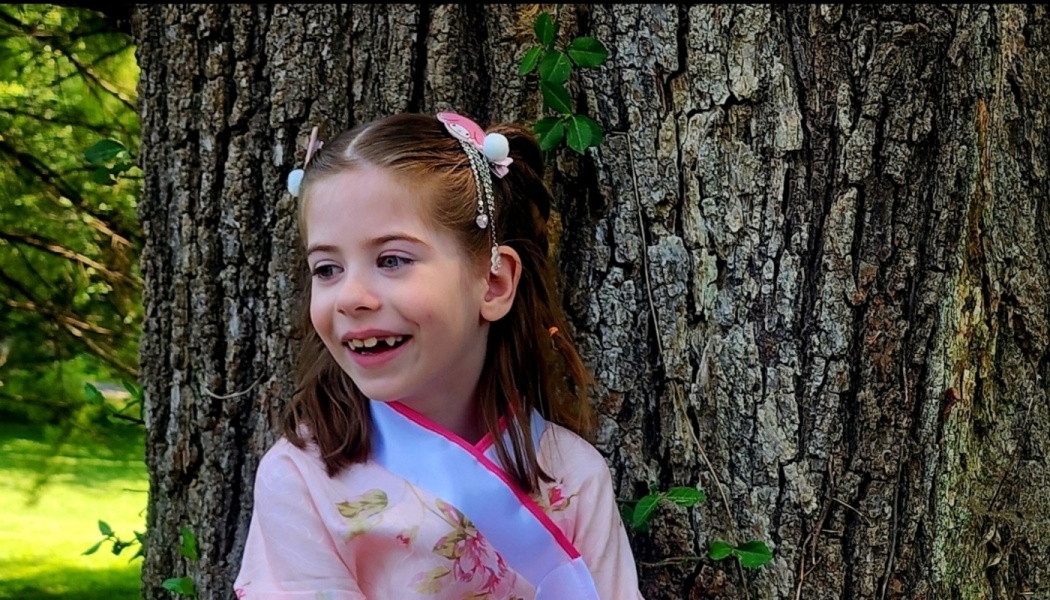 A smiling young girl in a pink floral traditional dress standing in front of a large tree outdoors.