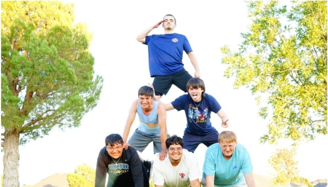 Six smiling young men form a three-tier human pyramid outdoors, with the person at the top saluting.