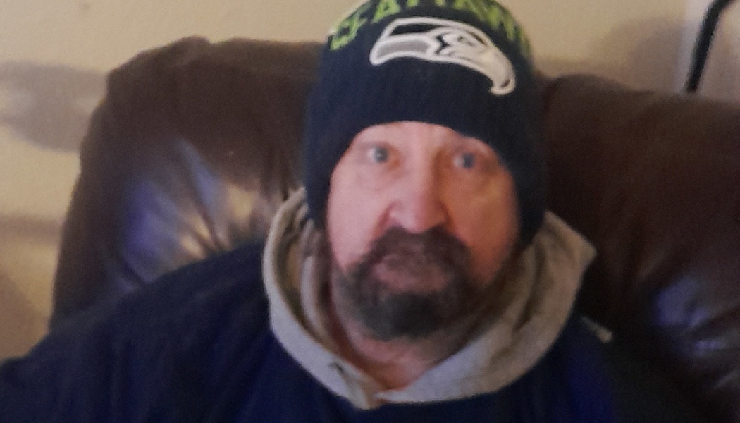 A bearded man wearing a Seattle Seahawks beanie and hoodie, sitting indoors on a brown leather chair.