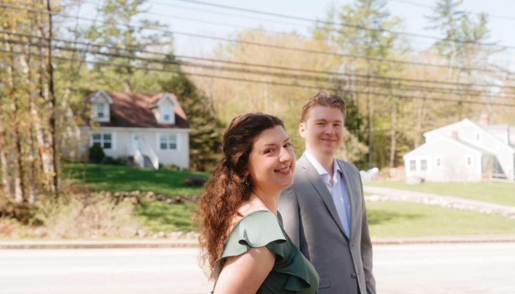 A smiling couple, a woman in a green dress and a man in a grey suit, standing outdoors in a suburban neighborhood.