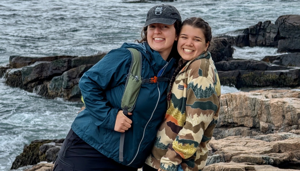Two women in outdoor jackets embrace and smile on a rocky coastline with ocean waves behind them.