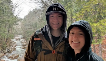 A smiling young couple wearing hooded jackets outdoors in the woods near a flowing stream.