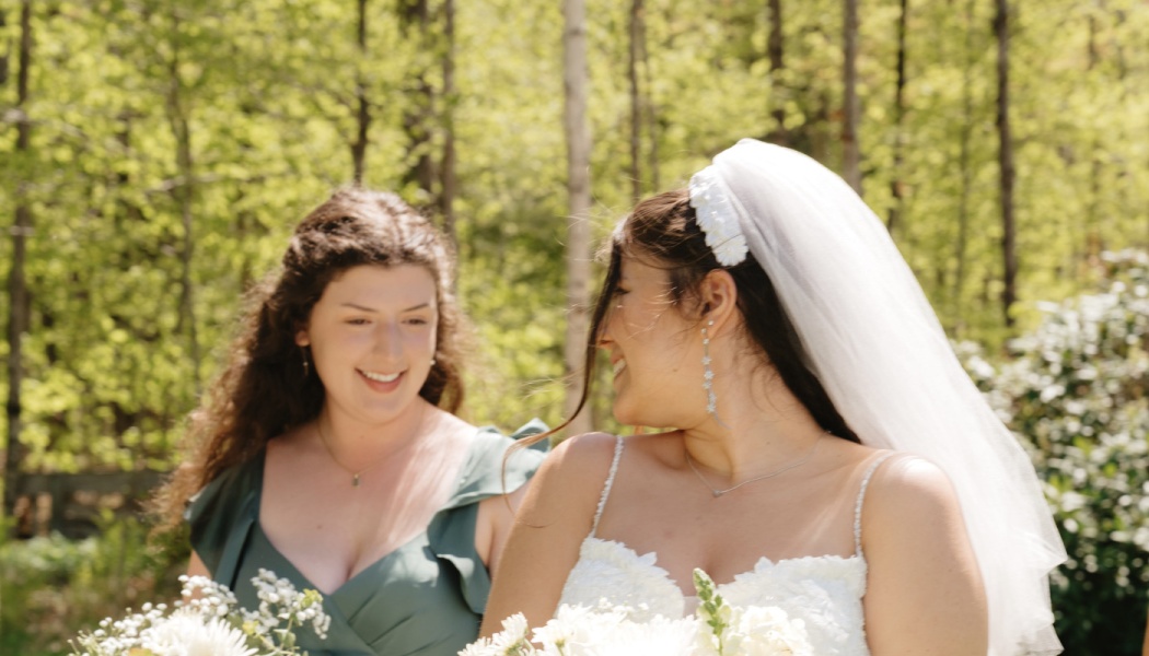 A smiling bride with a veil and a bridesmaid in green look at each other while holding white flower bouquets.