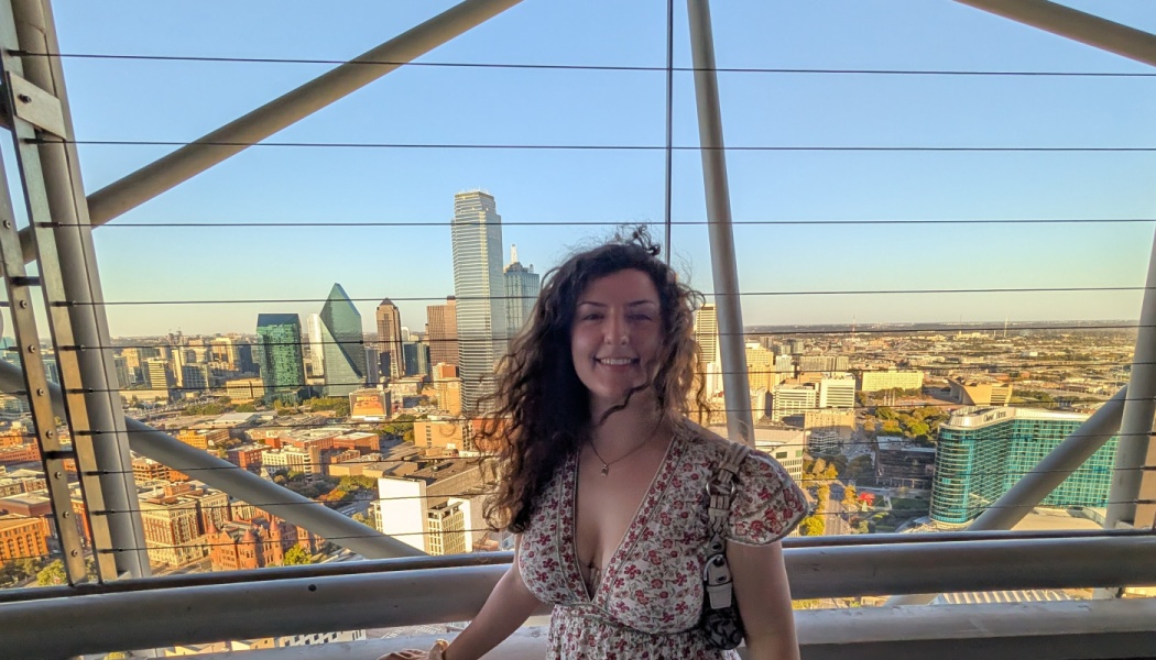 A smiling woman with long curly hair poses in a floral dress with a panoramic city skyline in the background.
