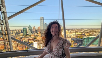A smiling woman with long curly hair poses in a floral dress with a panoramic city skyline in the background.
