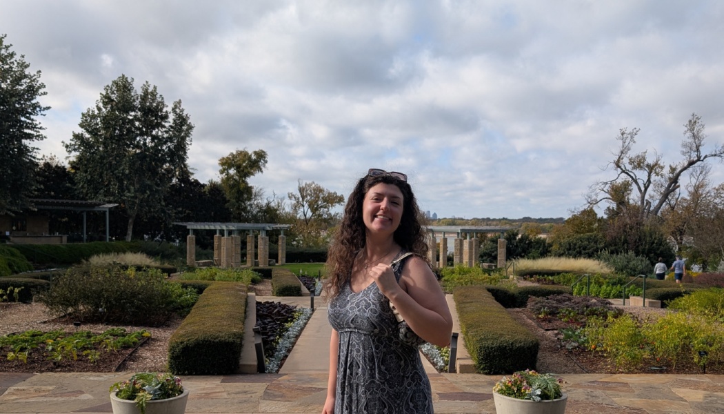 A smiling woman with long curly hair stands in a scenic garden with manicured hedges and stone structures.