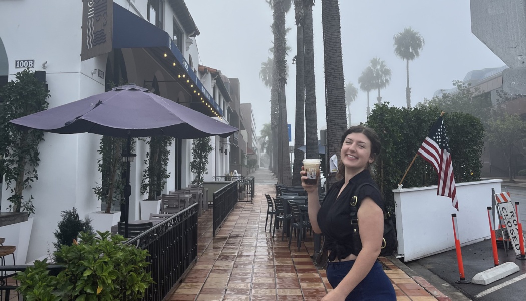 A smiling woman holds an iced coffee on a foggy street lined with palm trees and outdoor patio dining.