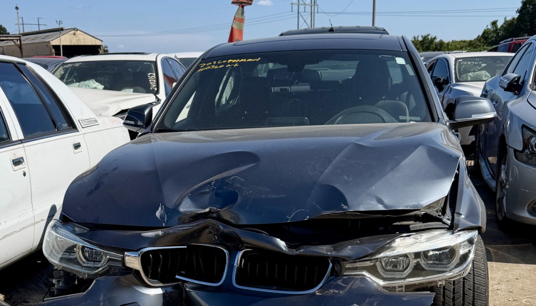 Dark grey BMW sedan with a severely crumpled hood and crushed front end in a salvage yard.
