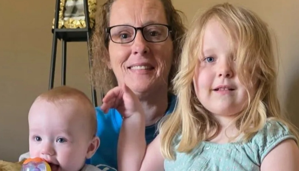 A smiling woman with glasses poses with a young blonde girl and a baby chewing on a toy.