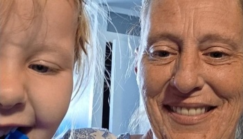 Close-up selfie of a smiling older woman next to a young child indoors.