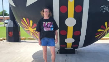 A woman in a black graphic t-shirt and denim shorts stands in front of a giant, colorful painted sphere sculpture.