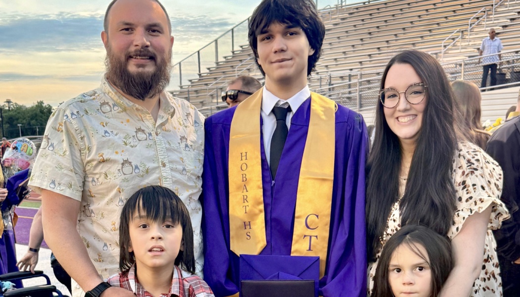 A proud family stands together at stadium bleachers to celebrate their graduate in a purple gown and gold sash.