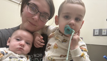 A mother wearing glasses poses closely with her baby and toddler holding a pacifier.