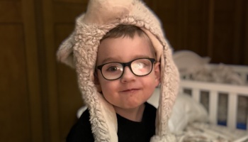 A young boy wearing black-framed glasses and a fluffy plush hat with ear flaps, smiling softly.