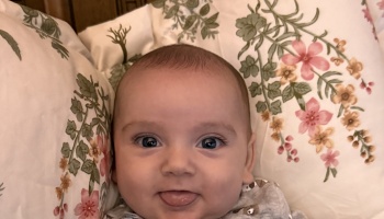 A smiling baby playfully sticking out their tongue while lying on floral patterned bedding.