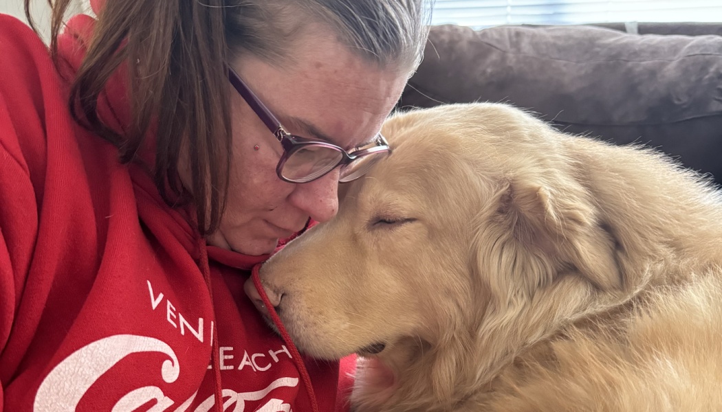 A woman in glasses and a red hoodie gently rests her forehead against a golden retriever with eyes closed.
