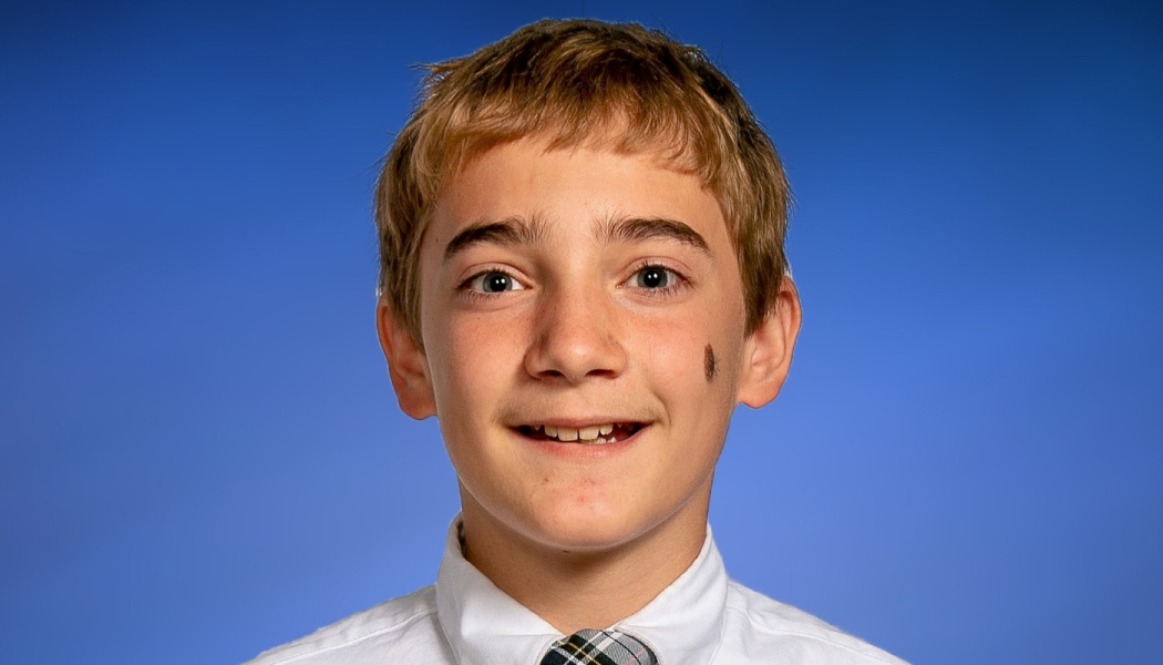 A smiling young boy with short light brown hair and a birthmark on his cheek, wearing a white shirt and tie.