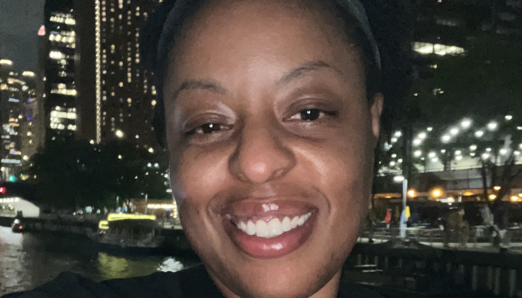 Close-up of a smiling Black woman outdoors at night with illuminated city lights reflecting on water behind her.