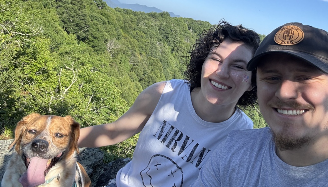 A smiling couple and their happy dog pose for a selfie on a scenic mountain overlook surrounded by trees.