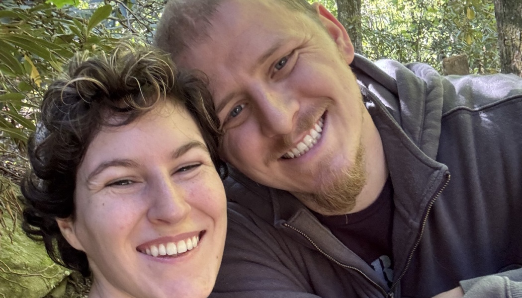 A smiling couple taking a close-up selfie outdoors surrounded by lush green trees.