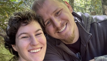 A smiling couple taking a close-up selfie outdoors surrounded by lush green trees.